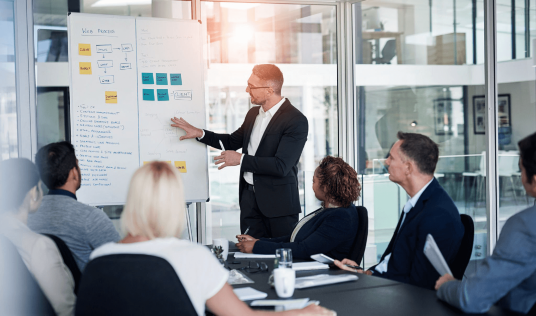 A presenter in a dark suit stands beside a whiteboard, gesturing towards a "Web Process" flowchart and other notes, while a group of people are seated around a conference table, attentively looking towards him during a business meeting. The room is brightly lit with a large window in the background.