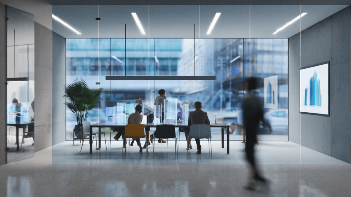 An expansive, modern office space features a large conference table with several people seated around it, looking towards a panoramic window that offers a view of city buildings. One person is blurred in the foreground, walking away from the viewer. The room has minimalist overhead lighting and a framed abstract blue artwork on the far wall. A large plant sits to the left of the table.
